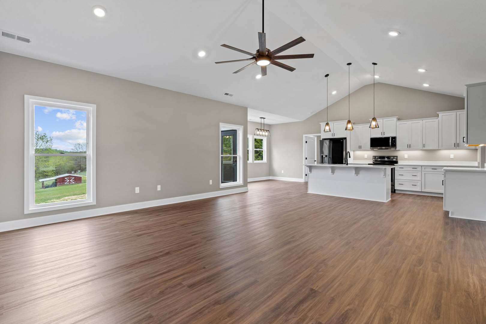 Spacious open floor plan featuring wood flooring, ceiling fan with light, large window overlooking a red barn and trees, and adjacent kitchen with cabinetry and appliances