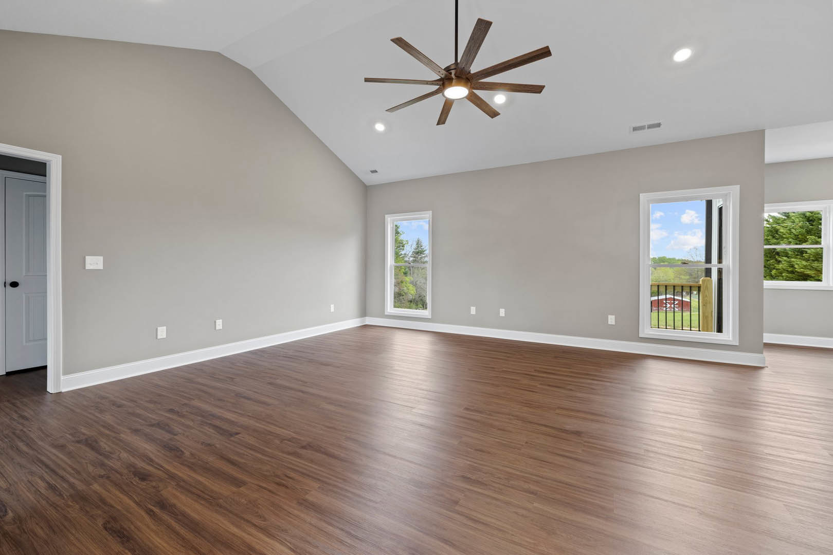 Spacious room with wood flooring, ceiling fan with lights, multiple windows showing trees and a barn outside, white door with black handle