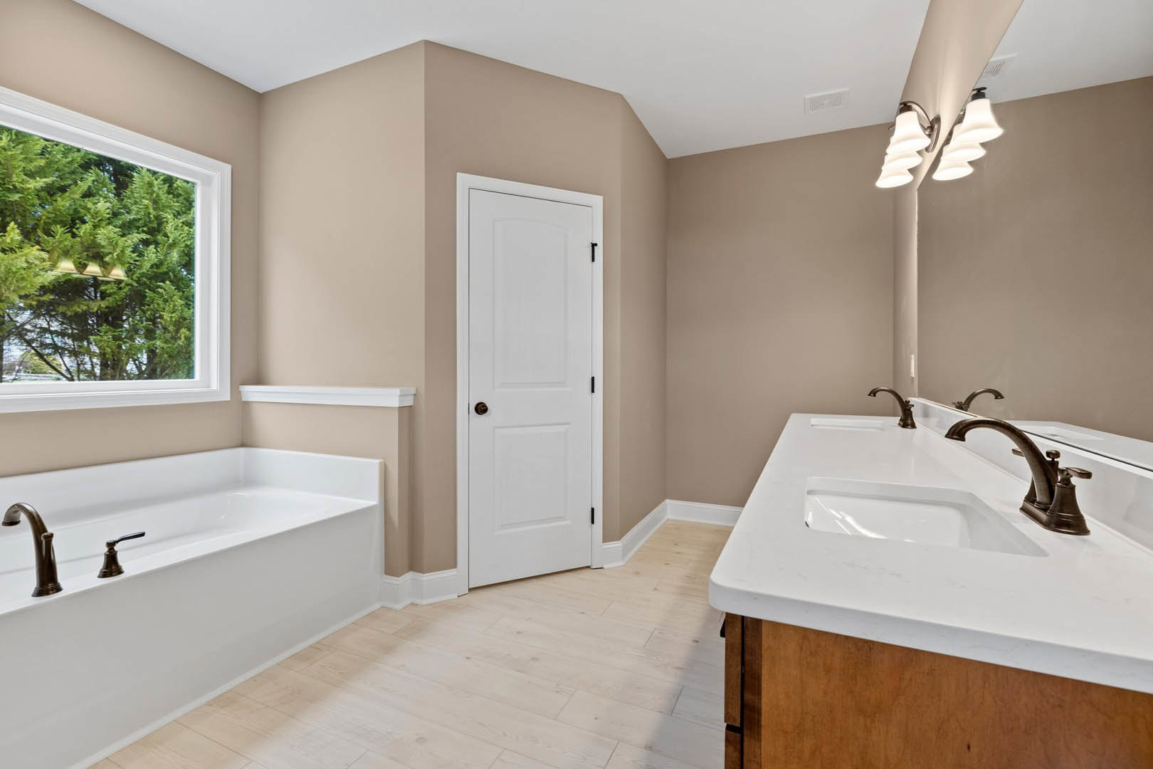 Bathtub with chrome faucets and wood flooring, white sink with brass faucet, white door with brass knob, window overlooking trees, modern light fixture on tiled wall