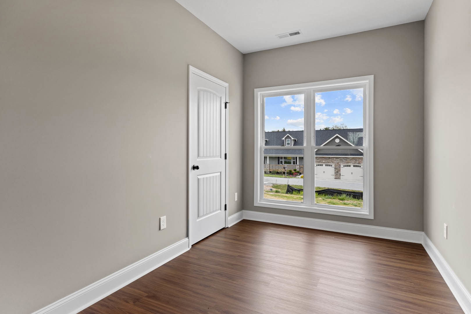 White paneled door and large window with blue sky view, wood flooring with white baseboard trim, interior wall, grassy yard visible outside.