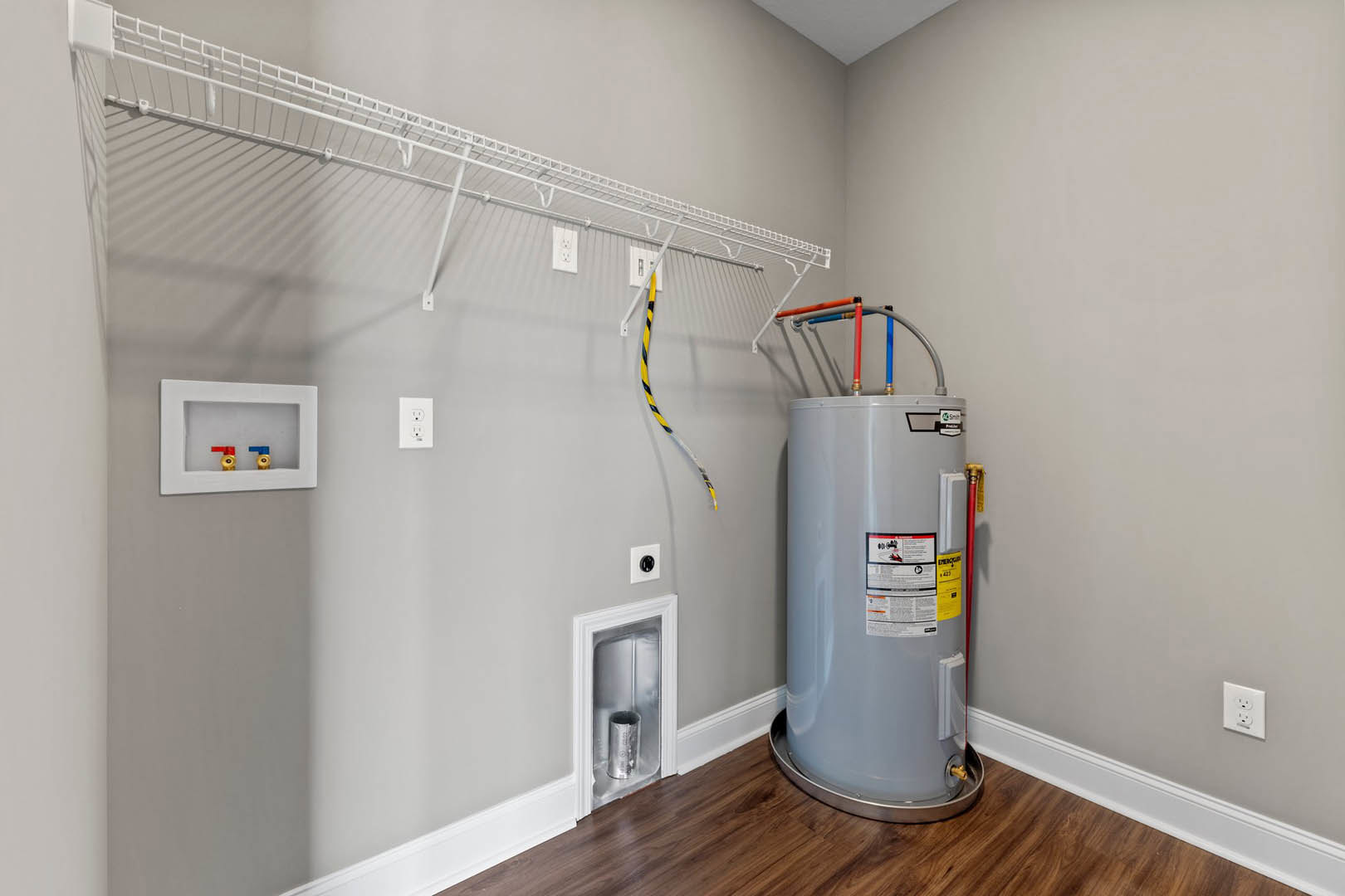 Utility room with white cylindrical water heater, visible valves, electrical outlets, silver ventilation tube near window, plaster walls, and light-colored flooring