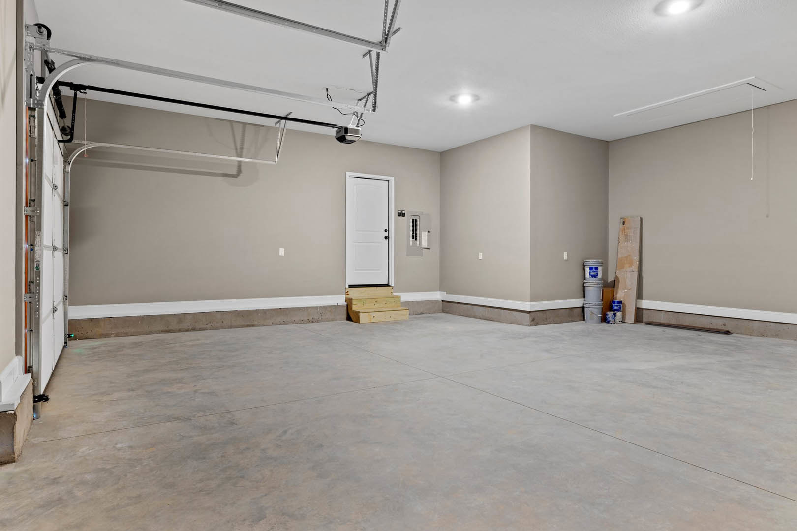 Spacious empty garage with concrete floor, white door featuring black knobs, wooden steps leading to interior, window above stairs, blue-labeled bucket near wall, overhead garage