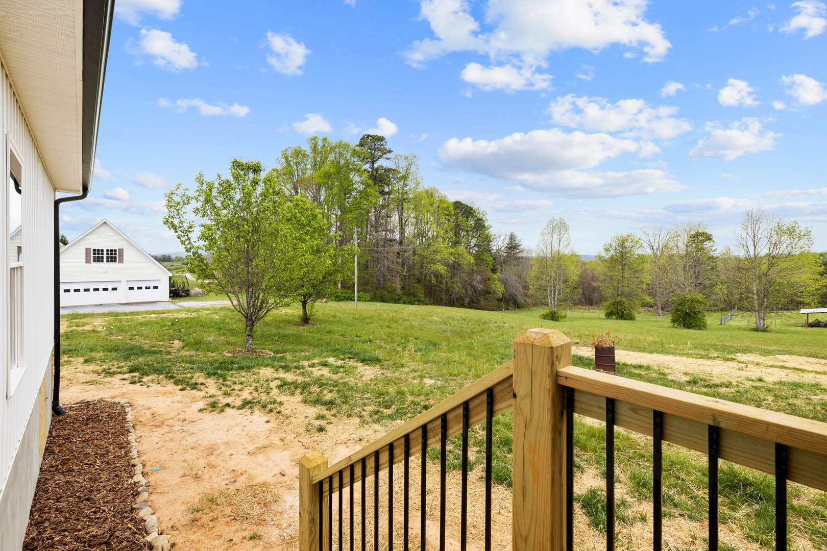 White house with attached garage set behind a wooden fence, surrounded by green grass, trees, and brown mulch; foreground features a wooden post and brown barrel with handle.