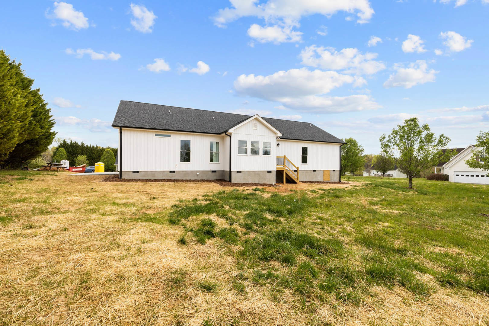 White farmhouse with black roof surrounded by grassy field, mature tree in foreground, blue sky with scattered clouds