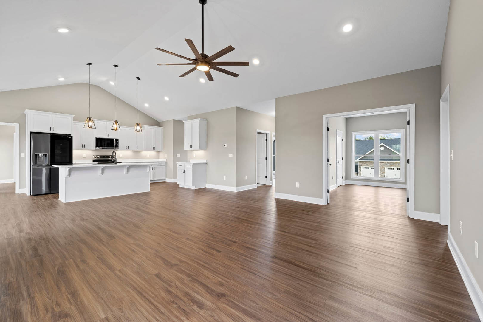 Spacious open floor plan featuring hardwood flooring, ceiling fan with light, stainless steel refrigerator, white walls with shelving, and large window overlooking neighboring
