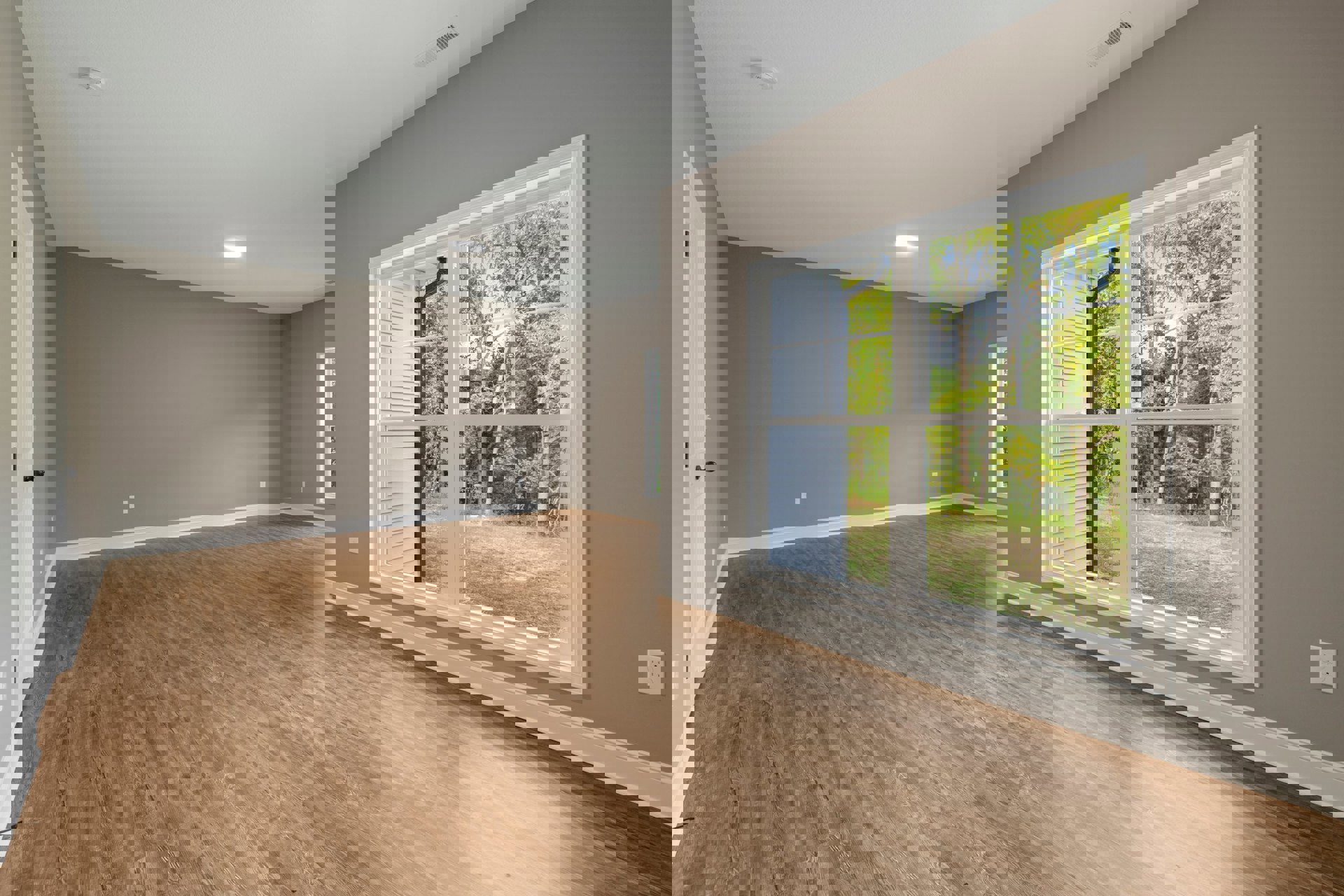 Sunlit room with wide window overlooking green trees, smooth hardwood floor, white walls, and a double electrical outlet.