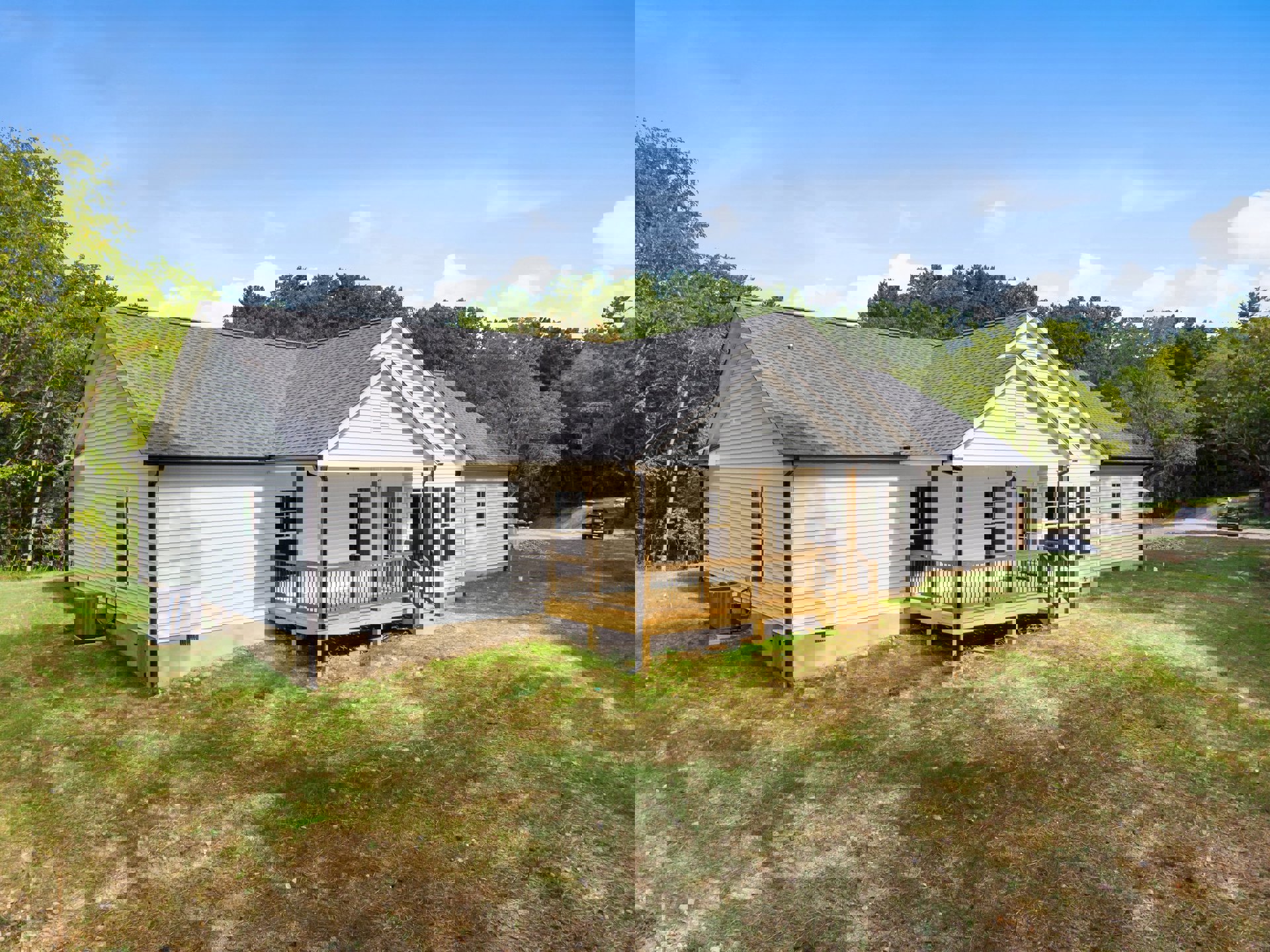 Two-story cottage with covered front porch, wooden deck, fenced lawn, heat pump unit, surrounded by trees under blue sky with scattered clouds