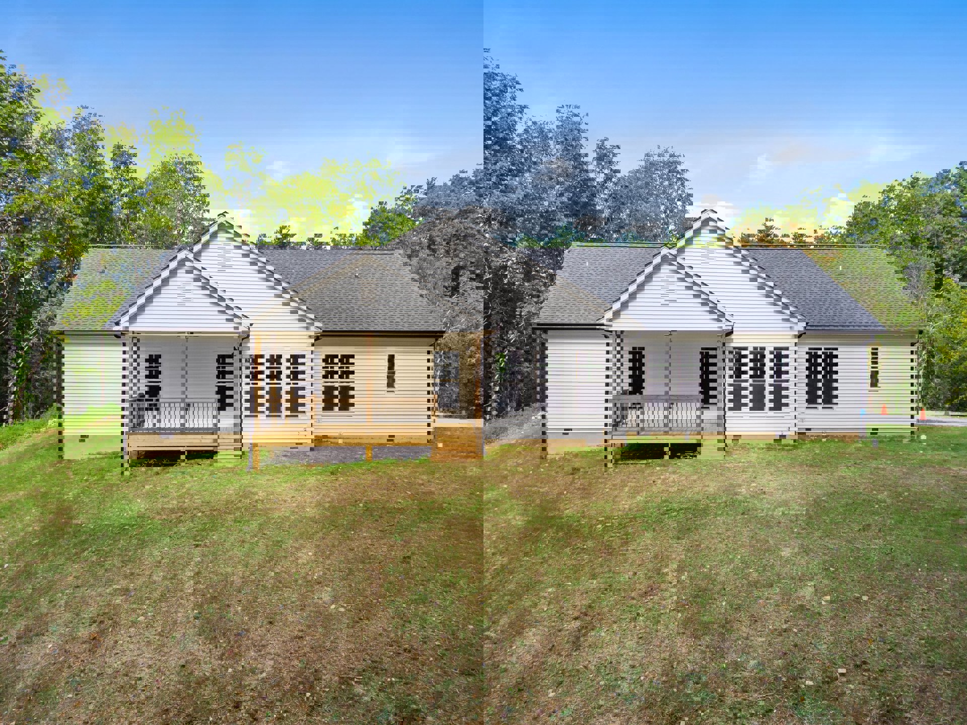 Two-story house with covered porch, white railing, wooden deck, manicured green lawn, large windows, gabled roof, and mature trees in the background