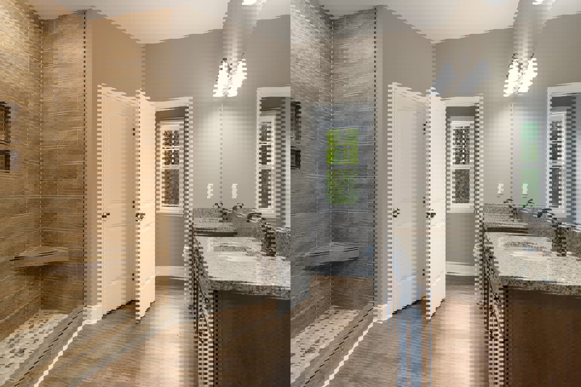 Bathroom with wood flooring and wood accent wall, granite countertops, white door with black knobs, corner shower, window overlooking trees.