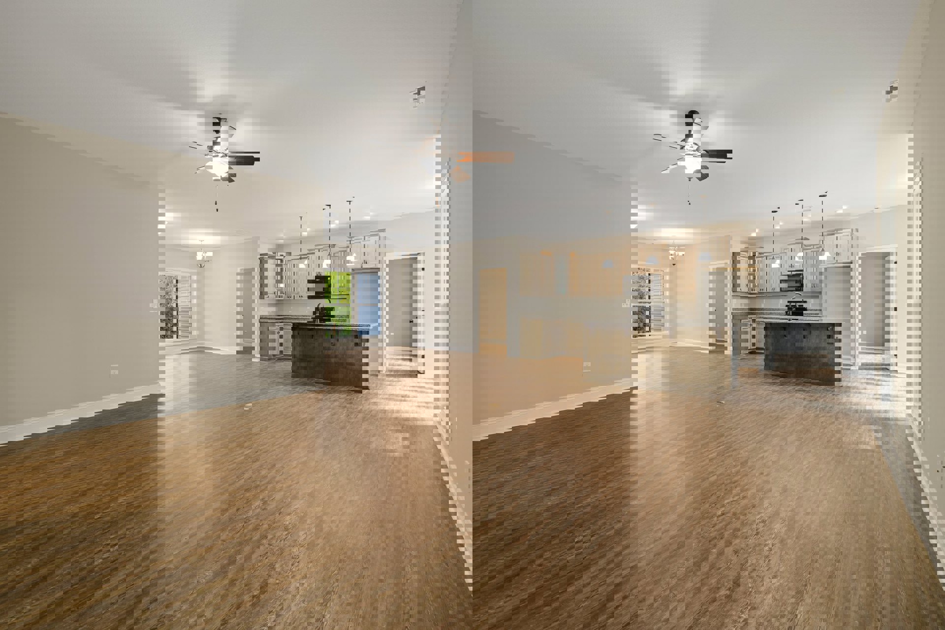 Spacious open floor plan featuring hardwood flooring, a central kitchen island with marble countertop, ceiling fan with light fixture, white door with black handles, and large