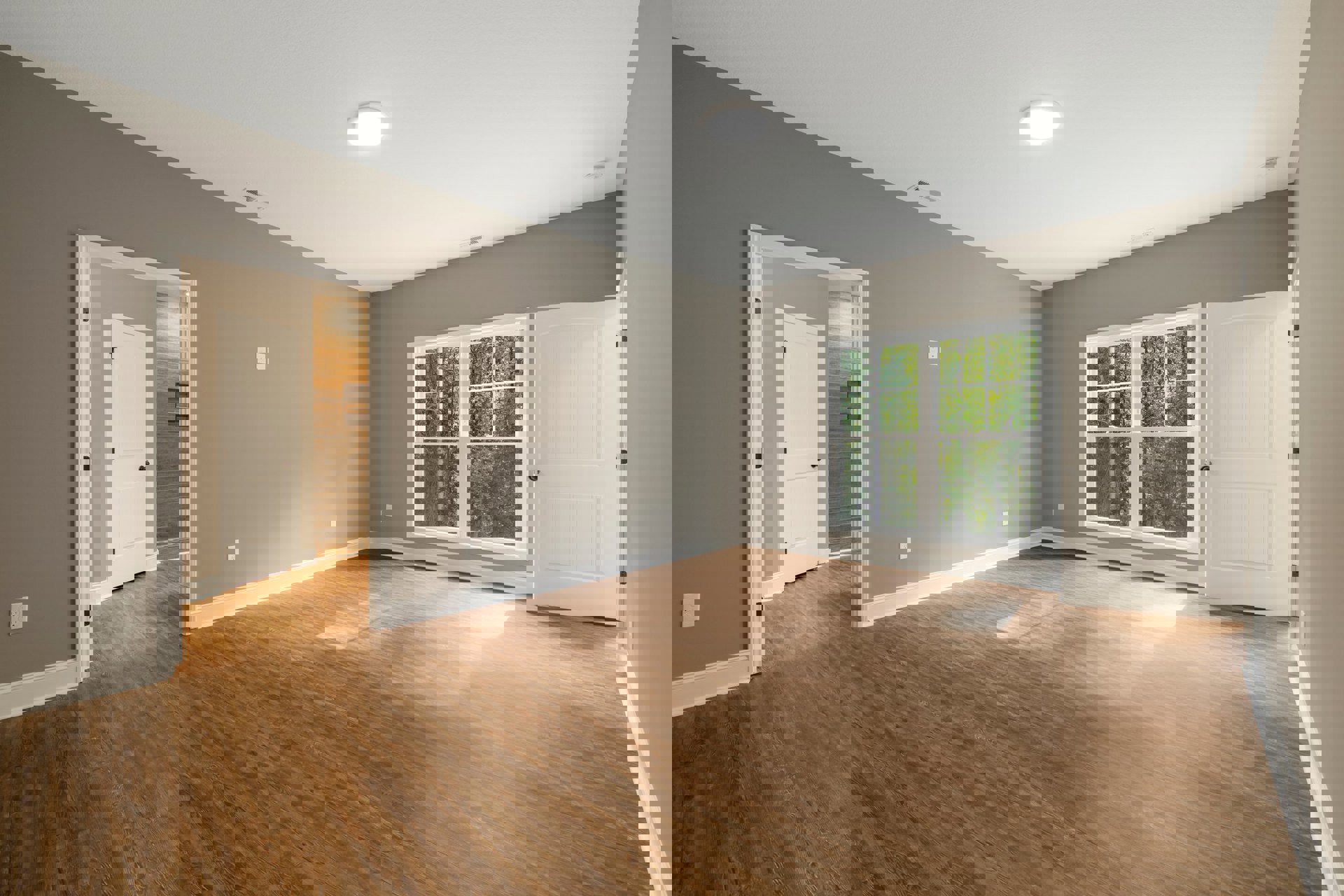 Wood flooring room with large window overlooking trees, white door with black hardware, ceiling light fixture, and neutral walls.