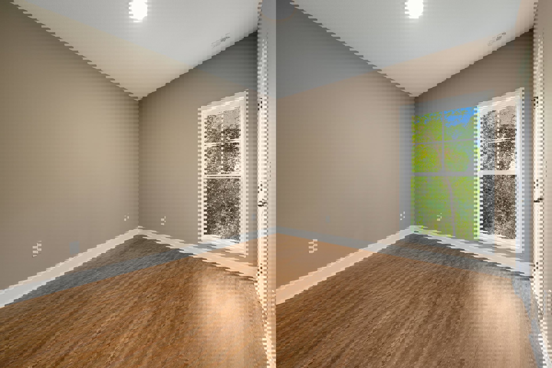 Hardwood floor with white trim, large window overlooking green trees, plaster walls, ceiling light fixture