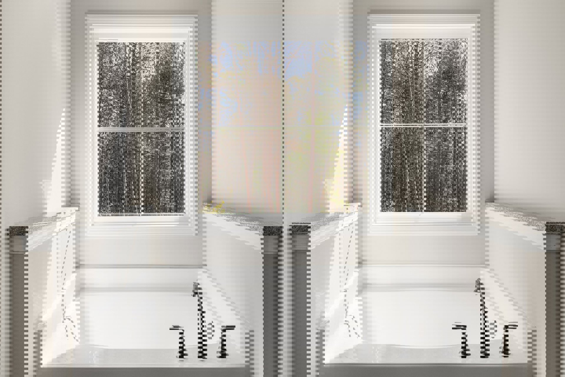 White freestanding bathtub beneath large window with leafy trees visible outside, white walls and shelf, chrome faucet, black bell-shaped decor on shelf