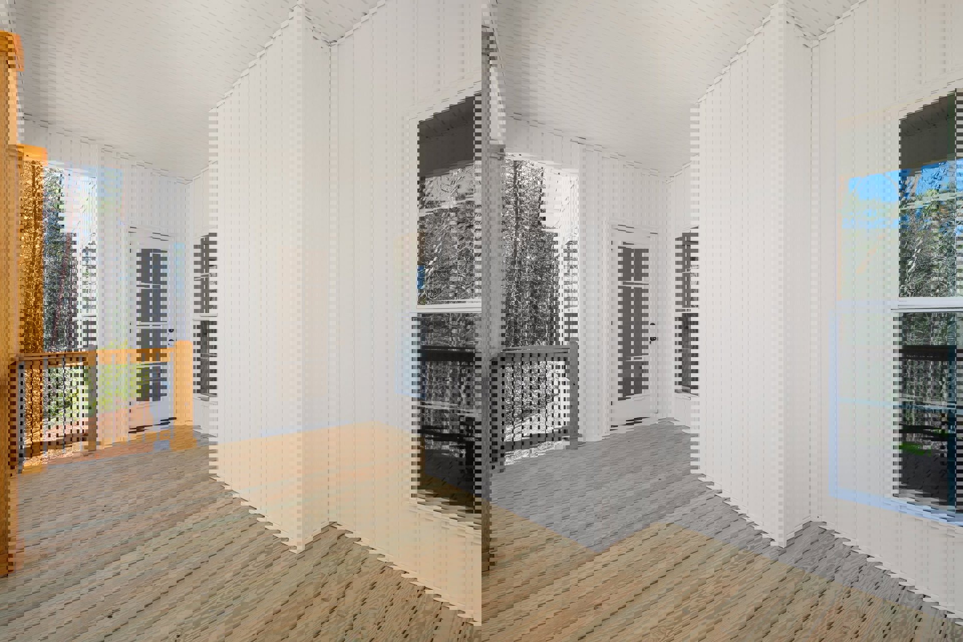 White-walled room with wood flooring, wooden railing featuring black bars, white door with glass panels, window overlooking trees, view of wooden deck with white siding through