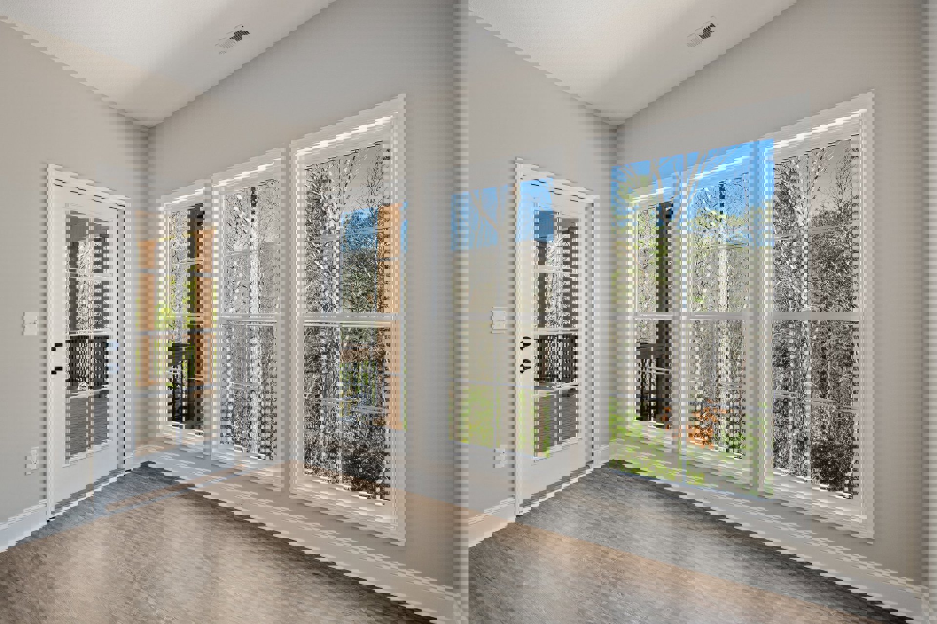 Bright room with white walls, wood laminate flooring, large windows overlooking trees, glass-paneled white door, and visible light switch on the wall.