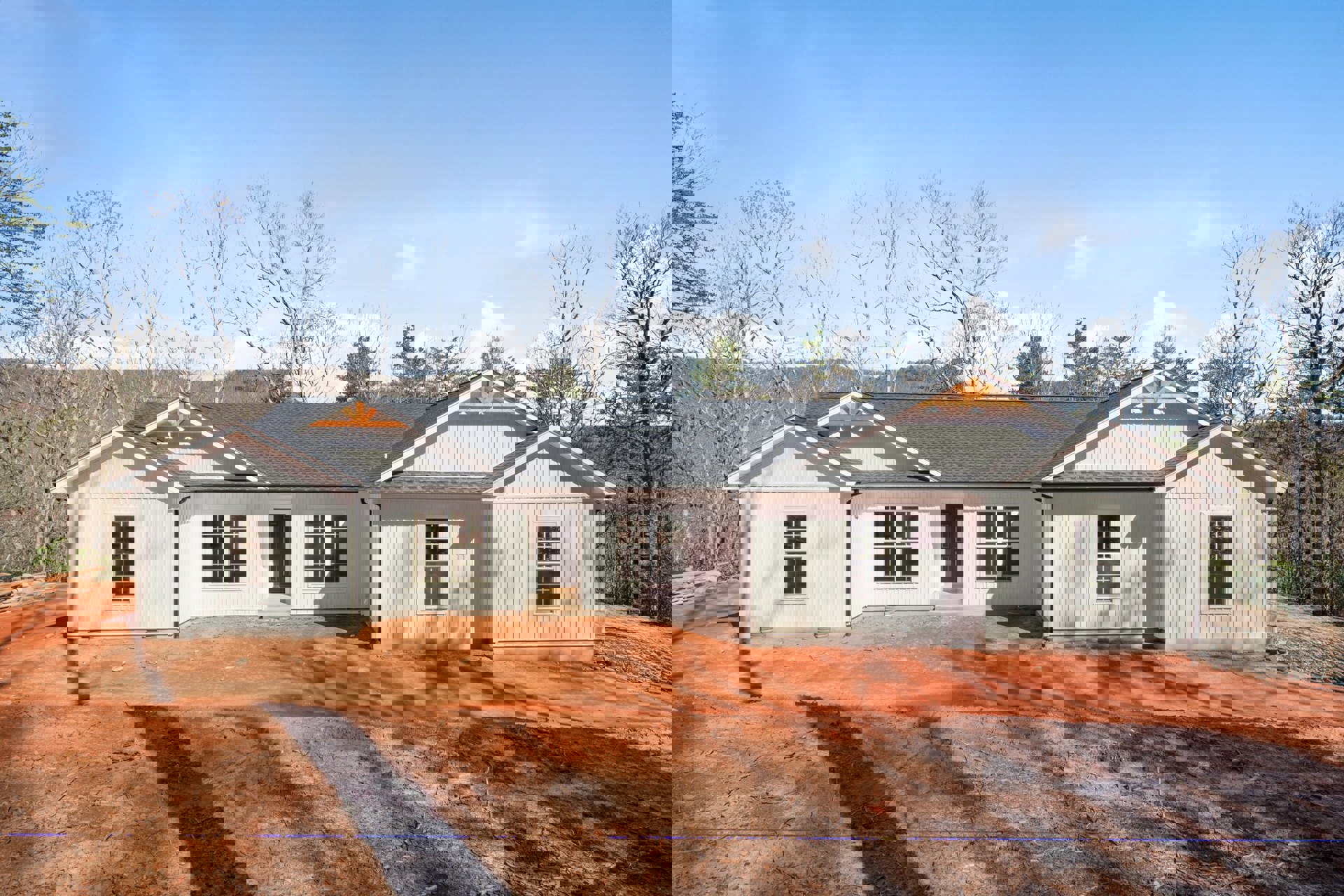 Partially built house with gray roof, black front door, white-framed windows, exposed dirt yard, leafless trees in background, cloudy sky overhead