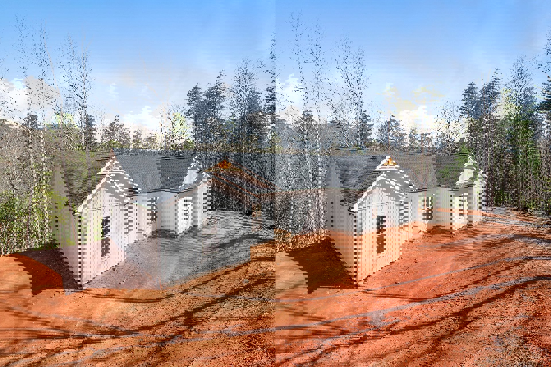 White house under construction surrounded by tall trees, dirt ground in foreground, new roof installed, blue sky with scattered clouds overhead