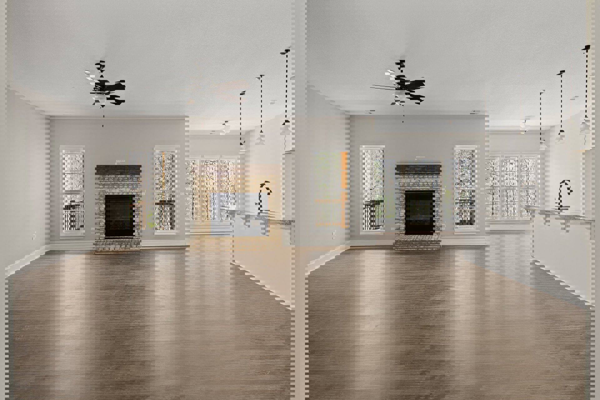 Spacious living room featuring wood flooring, central fireplace with black frame, ceiling fan with light fixture, plaster walls, and large window overlooking trees