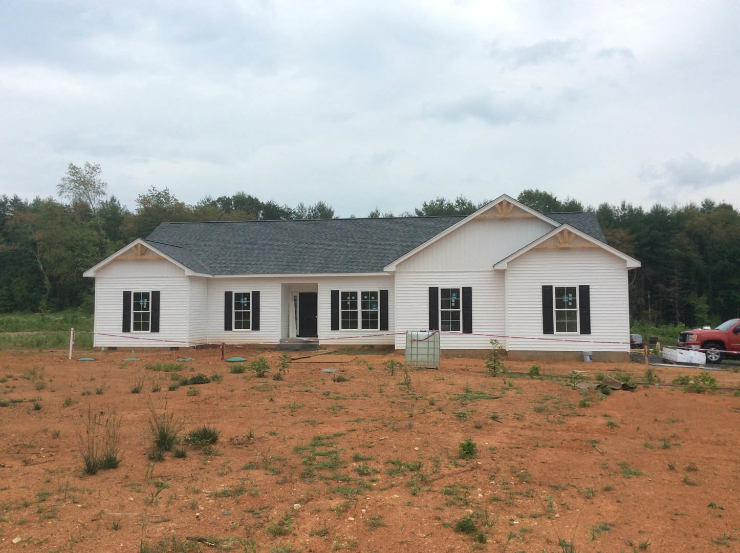 Two-story white house with black shutters under construction, surrounded by trees, dirt field with scattered plants, red truck parked nearby, cloudy sky overhead, white container