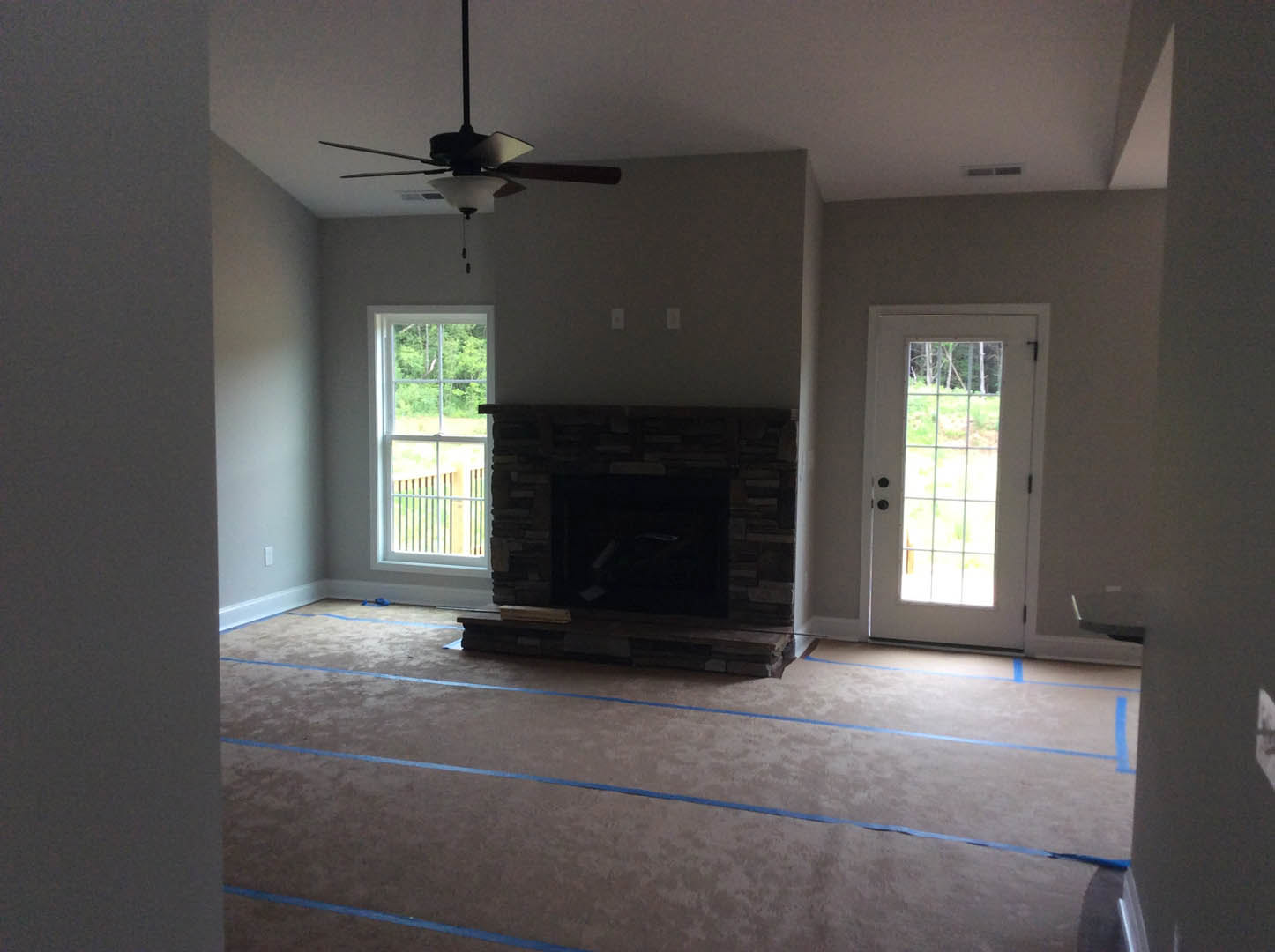 Living room with stone fireplace, ceiling fan with light, wood flooring marked with blue tape, windowed door, and large window showing outdoor view