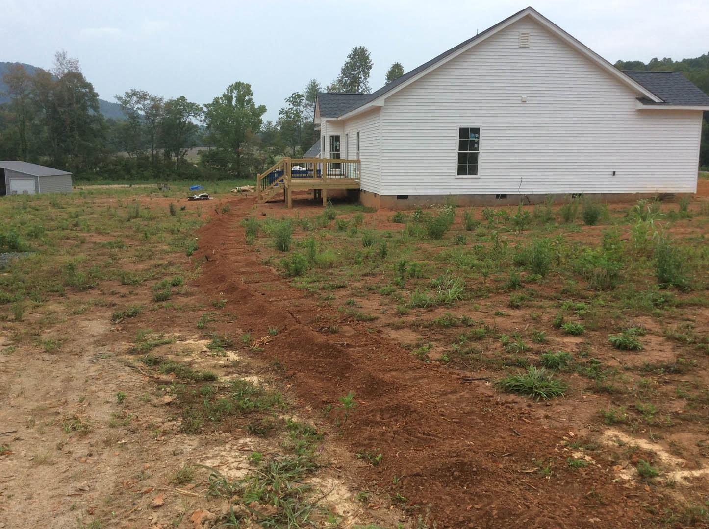 White cottage-style home with a wooden deck and railing, white-framed windows, fenced yard, dirt pathway leading to entrance, garage with white door, surrounded by grass, trees