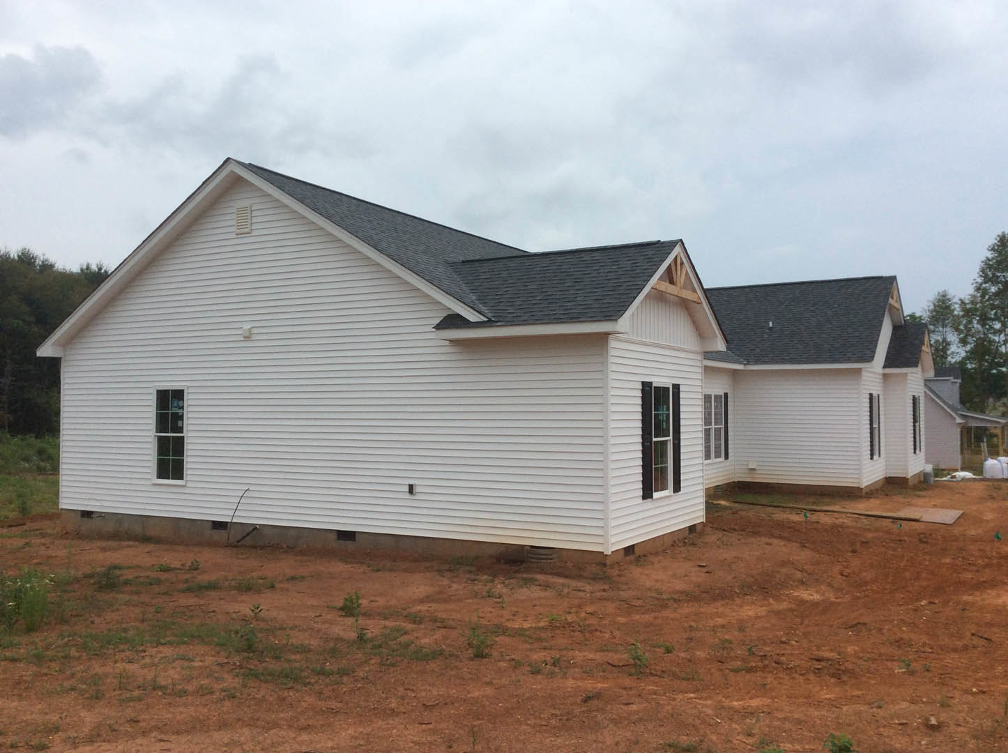 White siding house under construction with white-framed windows, unfinished yard with dirt and grass, and close-up view of roof against cloudy sky