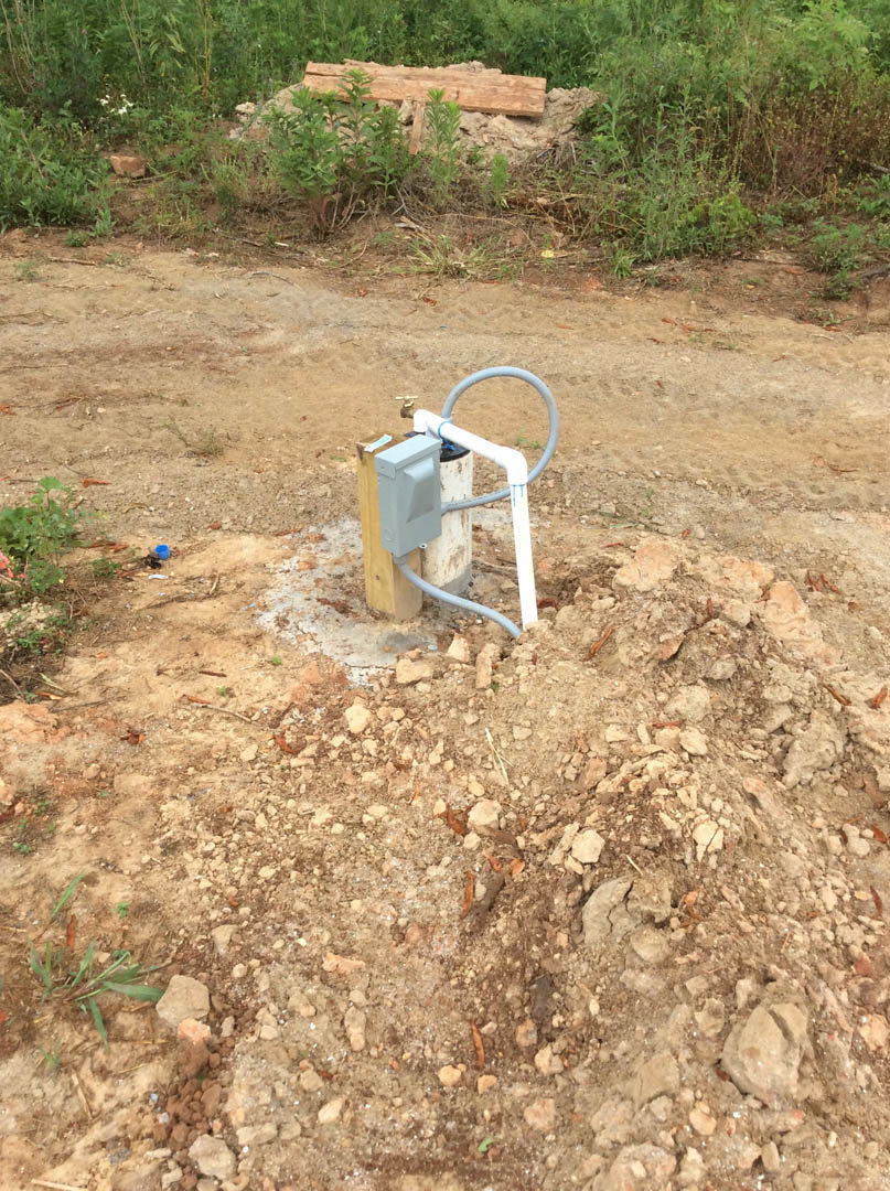 Water pump with attached pipe set in a patch of bare dirt, surrounded by grass and soil, with a rock wall and plants in the background