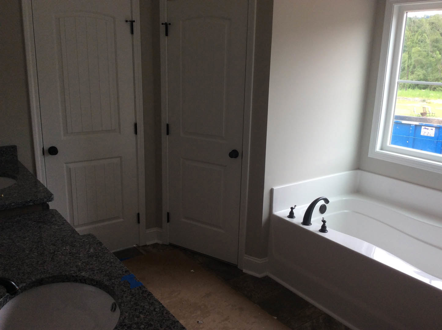White bathroom featuring a freestanding bathtub with black faucet, white sink with black hardware, white door with black knobs, large window overlooking lawn and outdoor chairs