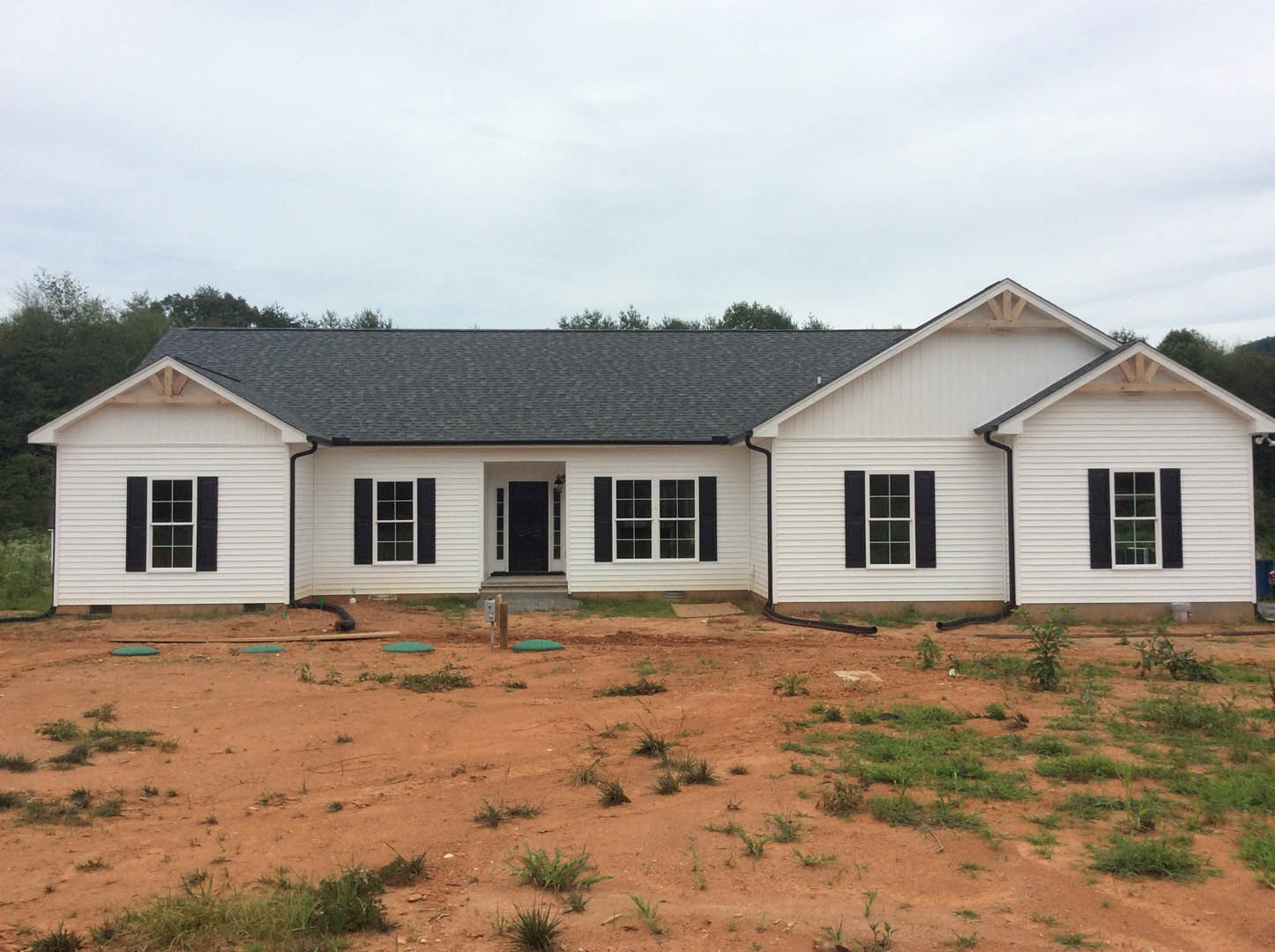 White siding house with black framed windows and black shutters, dirt yard with patches of grass, mature trees in background under partly cloudy sky