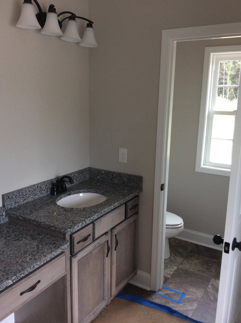 Bathroom with white sink and toilet, light fixture with white shade above, white door with window, blue tape on tiled floor, neutral walls, and cabinetry.
