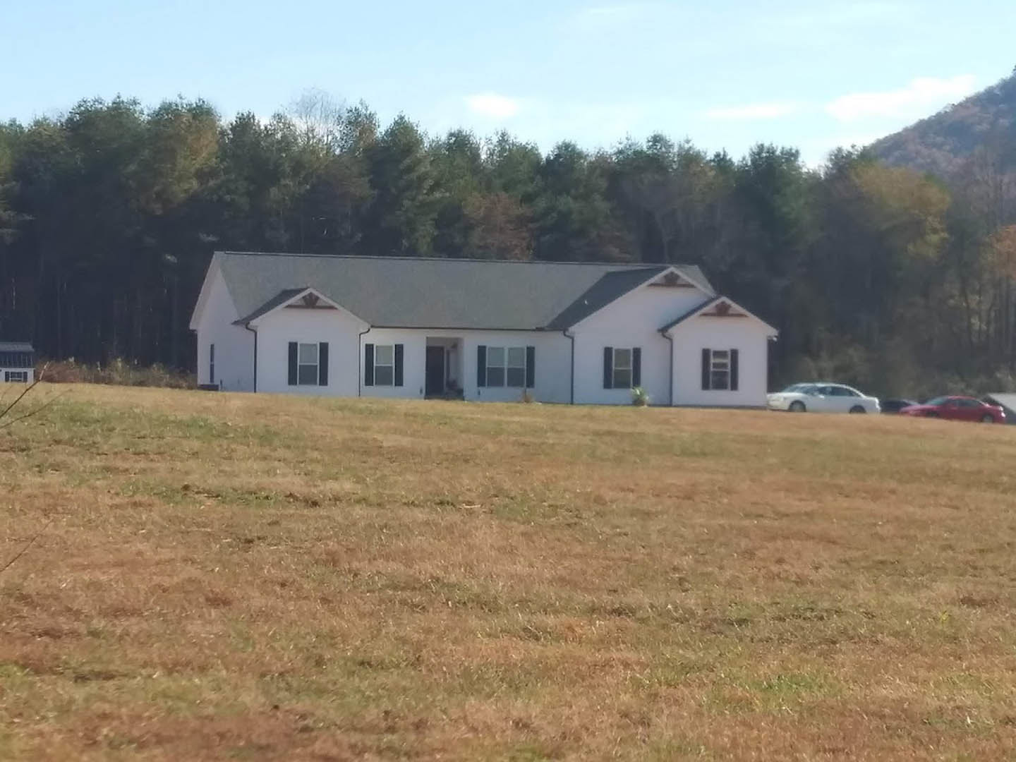 White farmhouse with black roof, parked car in driveway, grassy lawn, square-paned windows, trees and open sky in background