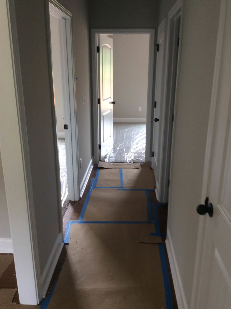 Hallway with laminate flooring, blue tape marking sections on the floor, white walls, plastic wrap near a white door, blurry black door knob, brown wood trim adjacent to white