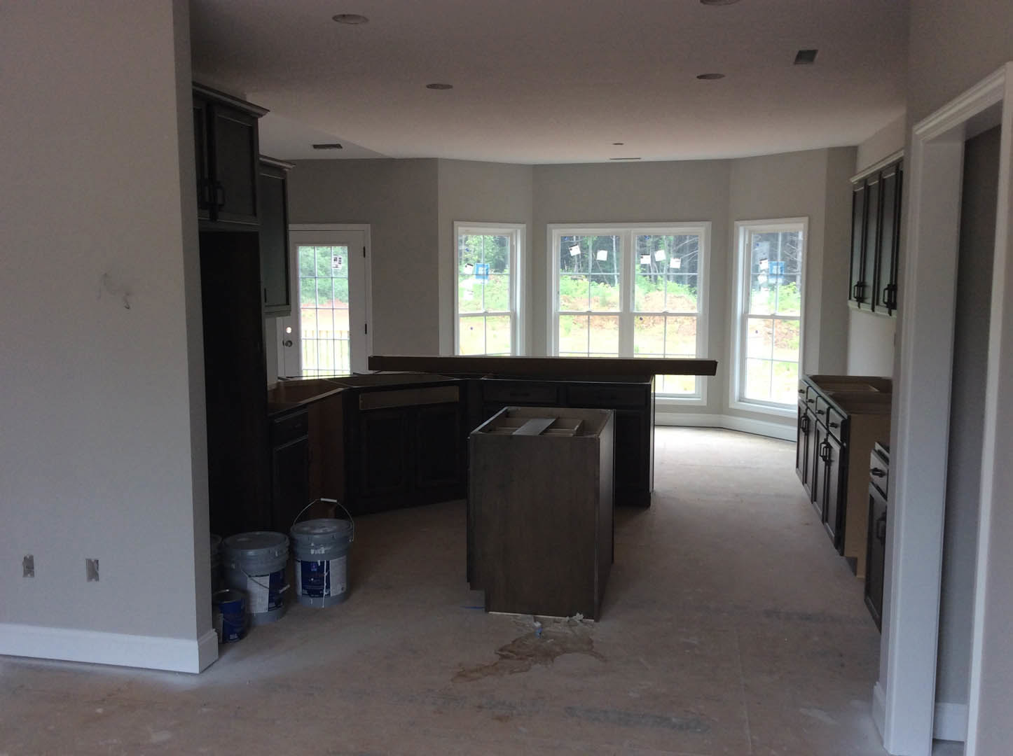 Modern kitchen with light wood cabinetry, dark stone countertops, stainless steel sink, and several plastic buckets placed on the floor; black accent wall and large window visible.