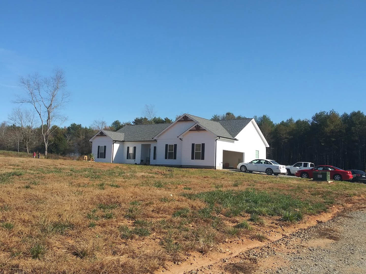 White two-story house with black shutters, leafless tree in front yard, grassy lawn, white and red cars parked along street.