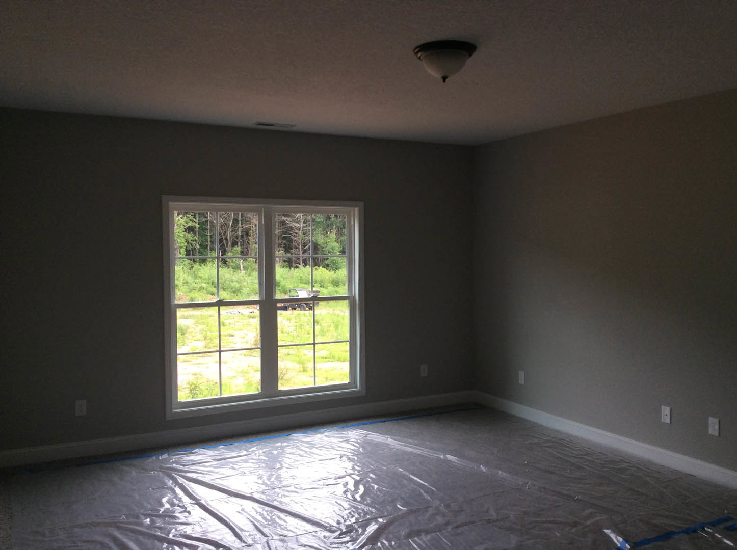 Bright room with large window overlooking field and trees, white plaster walls, plastic sheet covering hardwood floor, modern ceiling light fixture