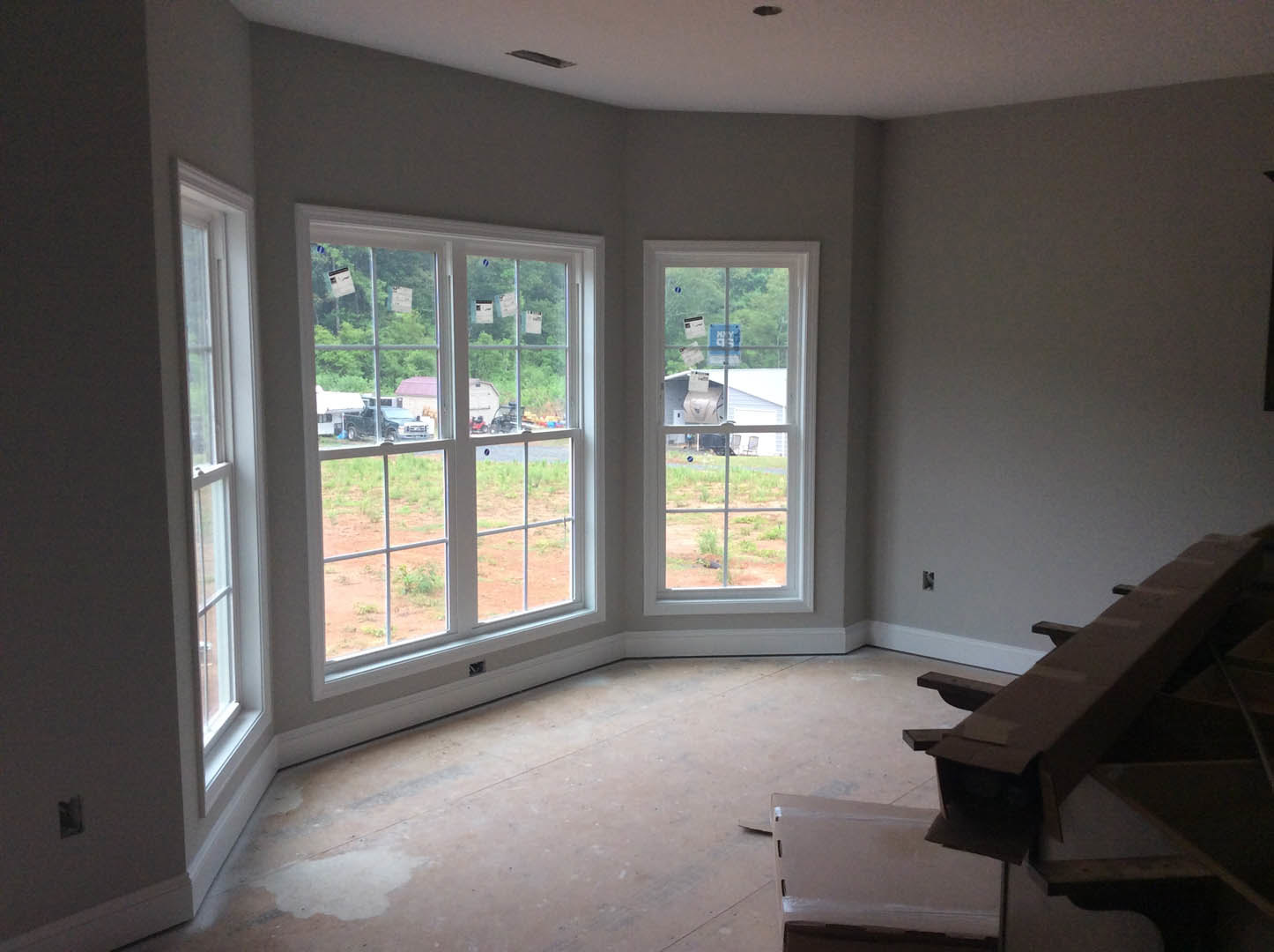 Dining area with large windows overlooking a field and trees, wooden table with a cardboard box, light-colored flooring, white walls, and a window sign visible.