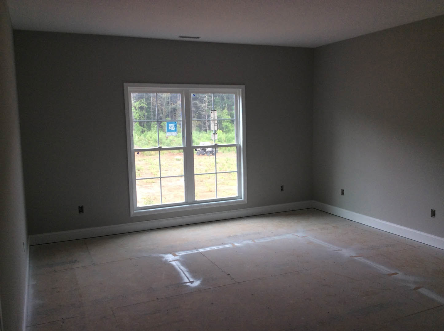 Concrete-floored room with plaster walls, large window, and white line marking on floor; tractor and tree visible outside through glass.