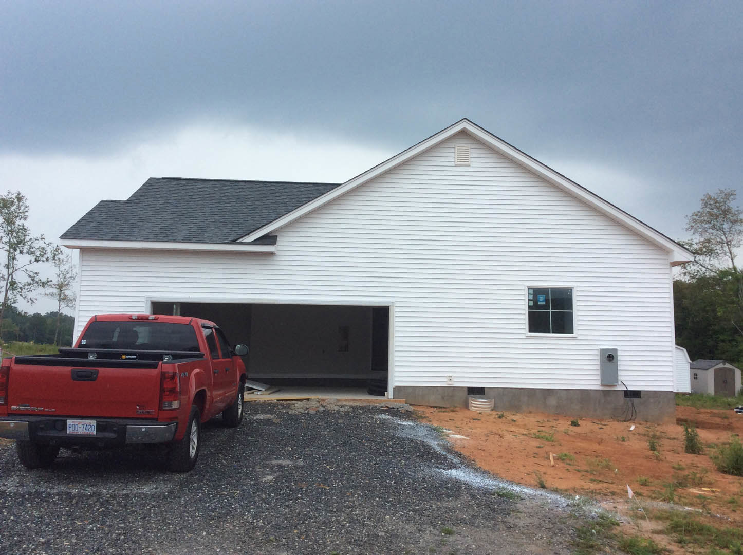 White house with horizontal siding and black-framed windows, red pickup truck parked on gravel driveway, leafy tree beside the house, gray shingle roof under partly cloudy sky