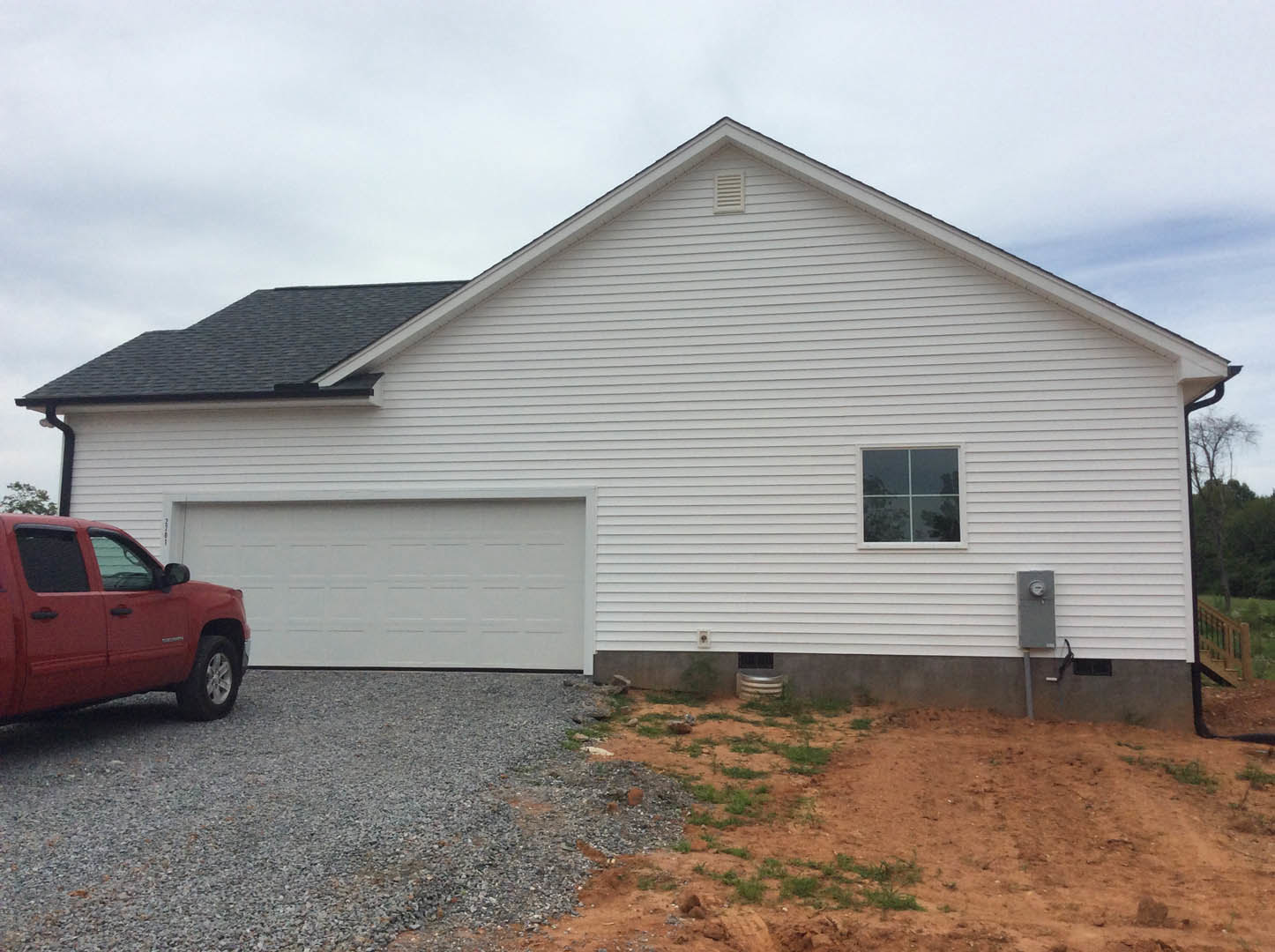 White siding house with attached garage, red pickup truck parked on gravel driveway, white-framed windows, patchy grass and dirt yard, partly cloudy sky overhead