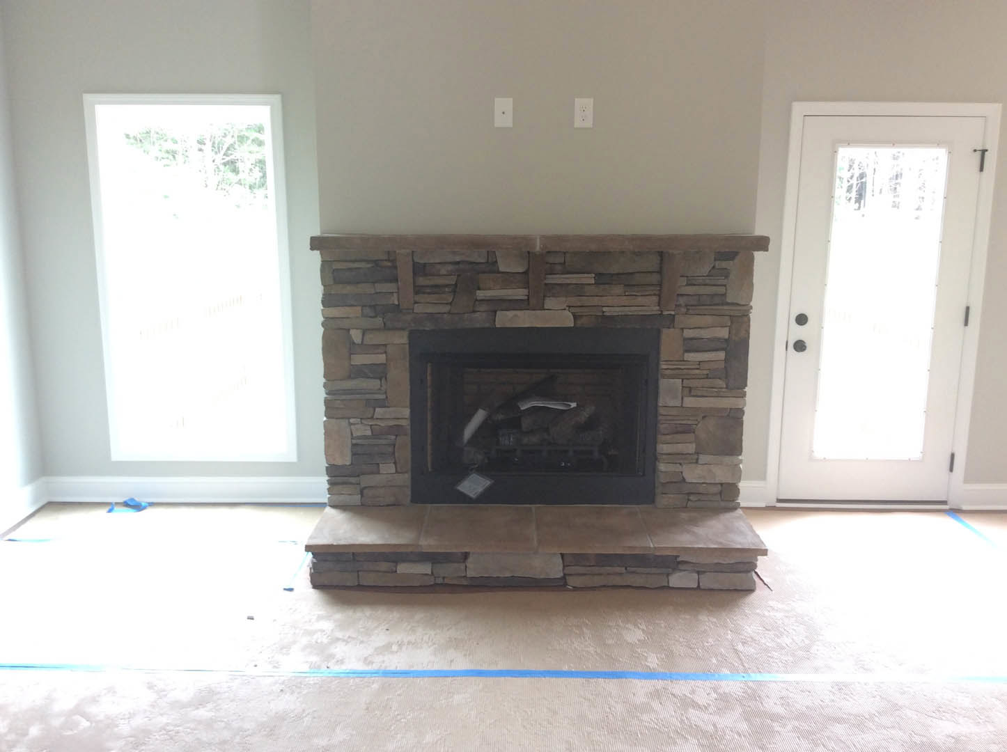 Stone fireplace with stacked logs, white door with window, hardwood flooring, and sunlight streaming through large window in cozy living room.