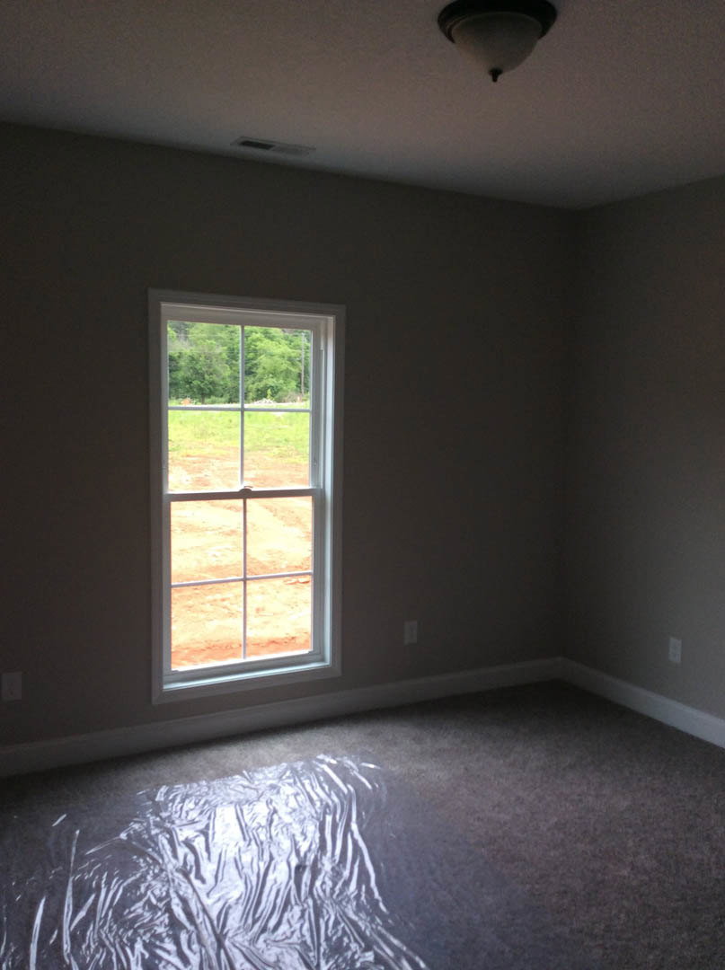 Sunlit room with plaster walls, large window overlooking dirt field, light fixture on ceiling, plastic wrap and bag on unfinished floor