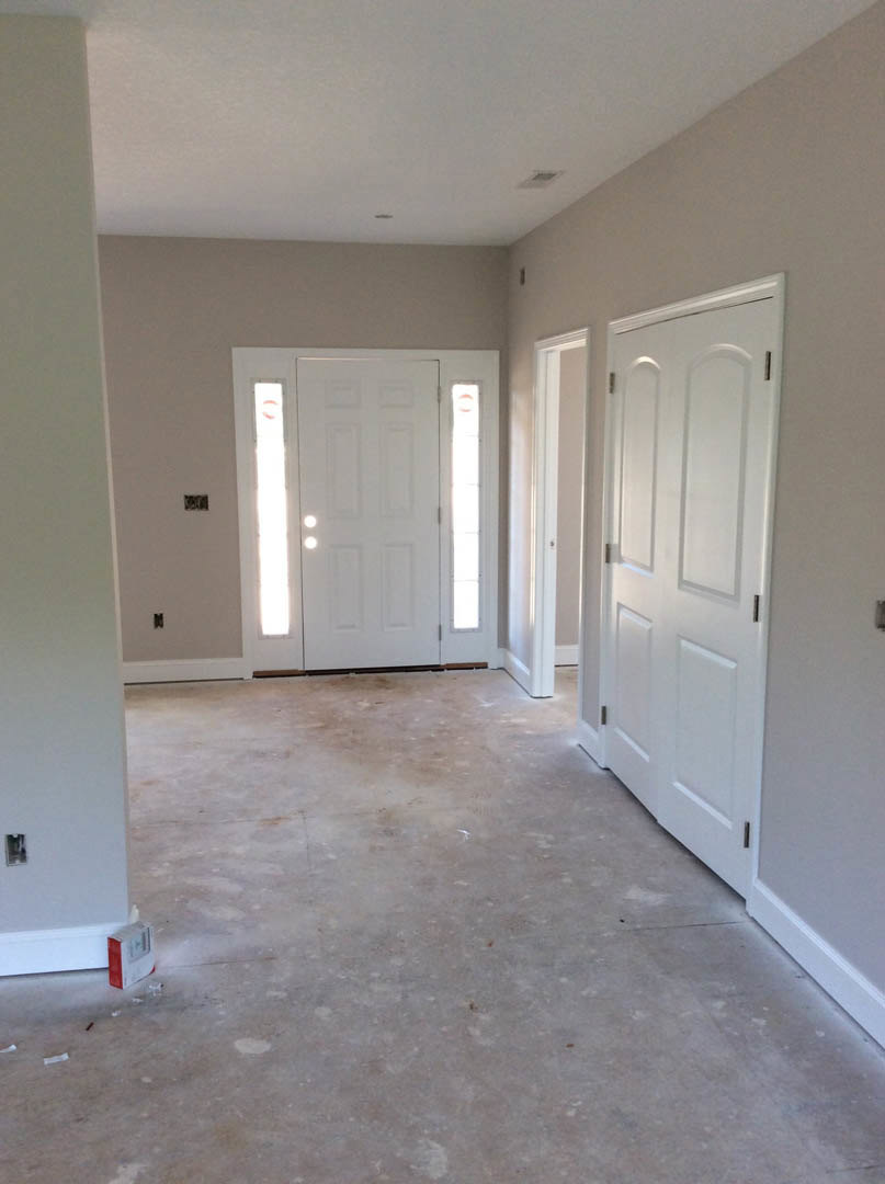 Hallway with white paneled doors, white trim, and light-colored flooring; white door features two glass windows and a matching door frame; walls finished in smooth white plaster.