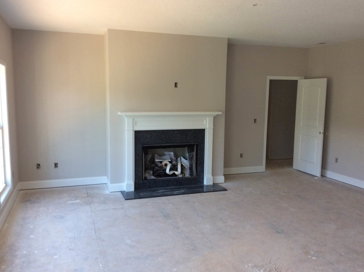 Stone fireplace with black hearth, white plaster walls, hardwood flooring, white door with silver knob, roll of toilet paper placed in fireplace opening, sunlight reflecting on