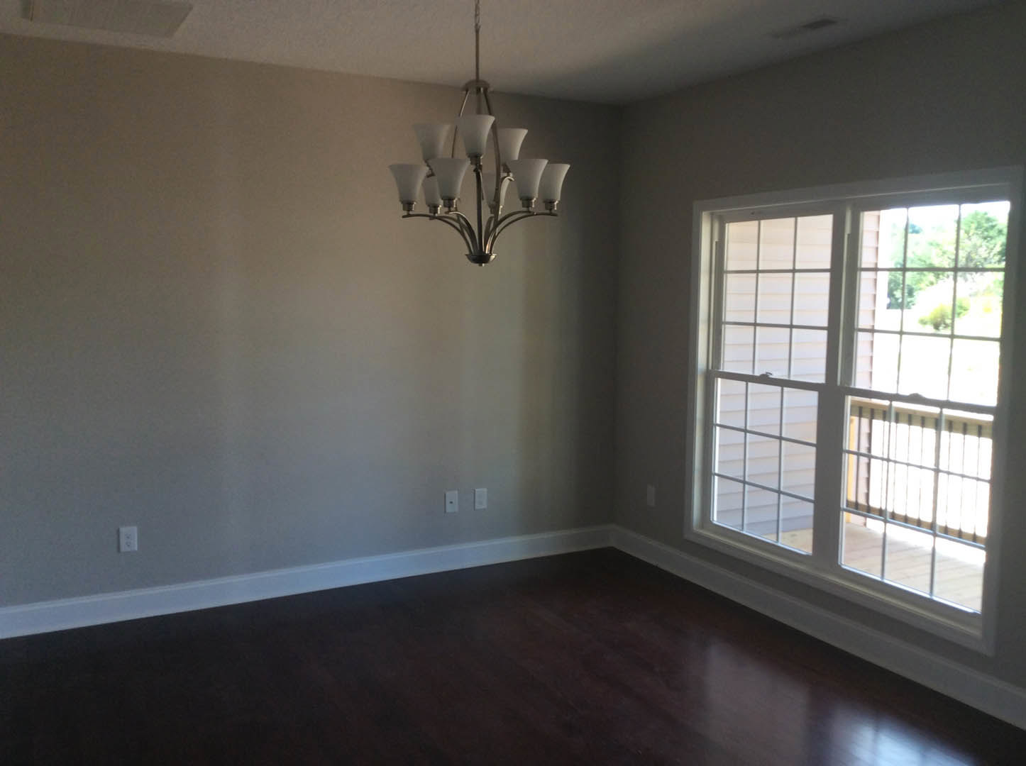 Dark wood floor with white baseboard, large window overlooking deck with railing, chandelier featuring white shades, white rectangular outlet on wall