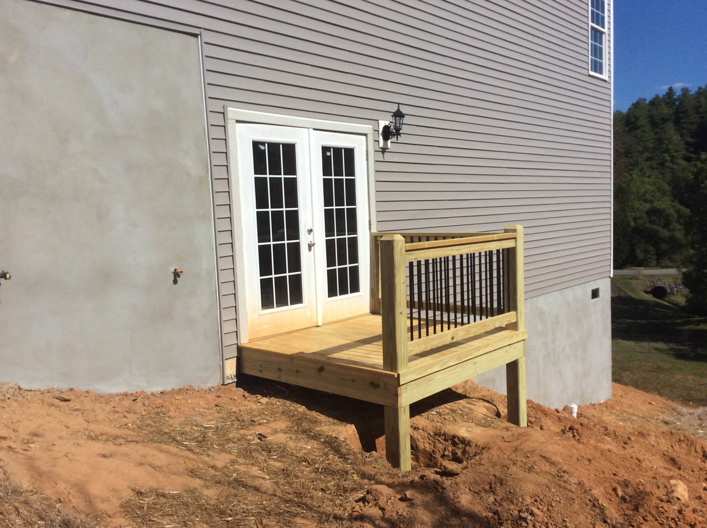 Wooden deck with metal railing attached to a house, white double doors with glass panes, exterior wall with visible pipes, forested background.
