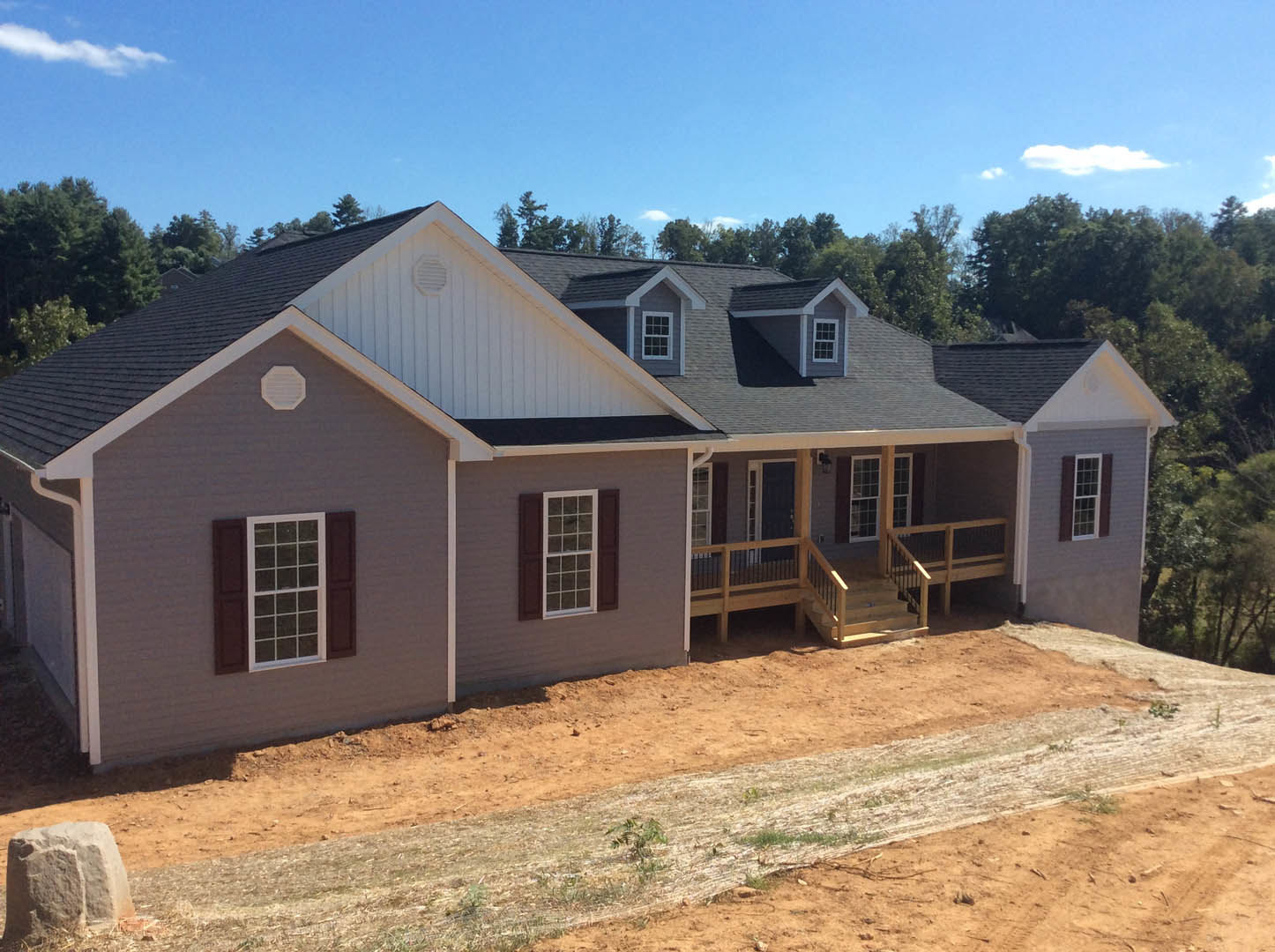 White custom home with black trim, large windows, and dirt road leading to entrance; trees and cloudy sky in background.