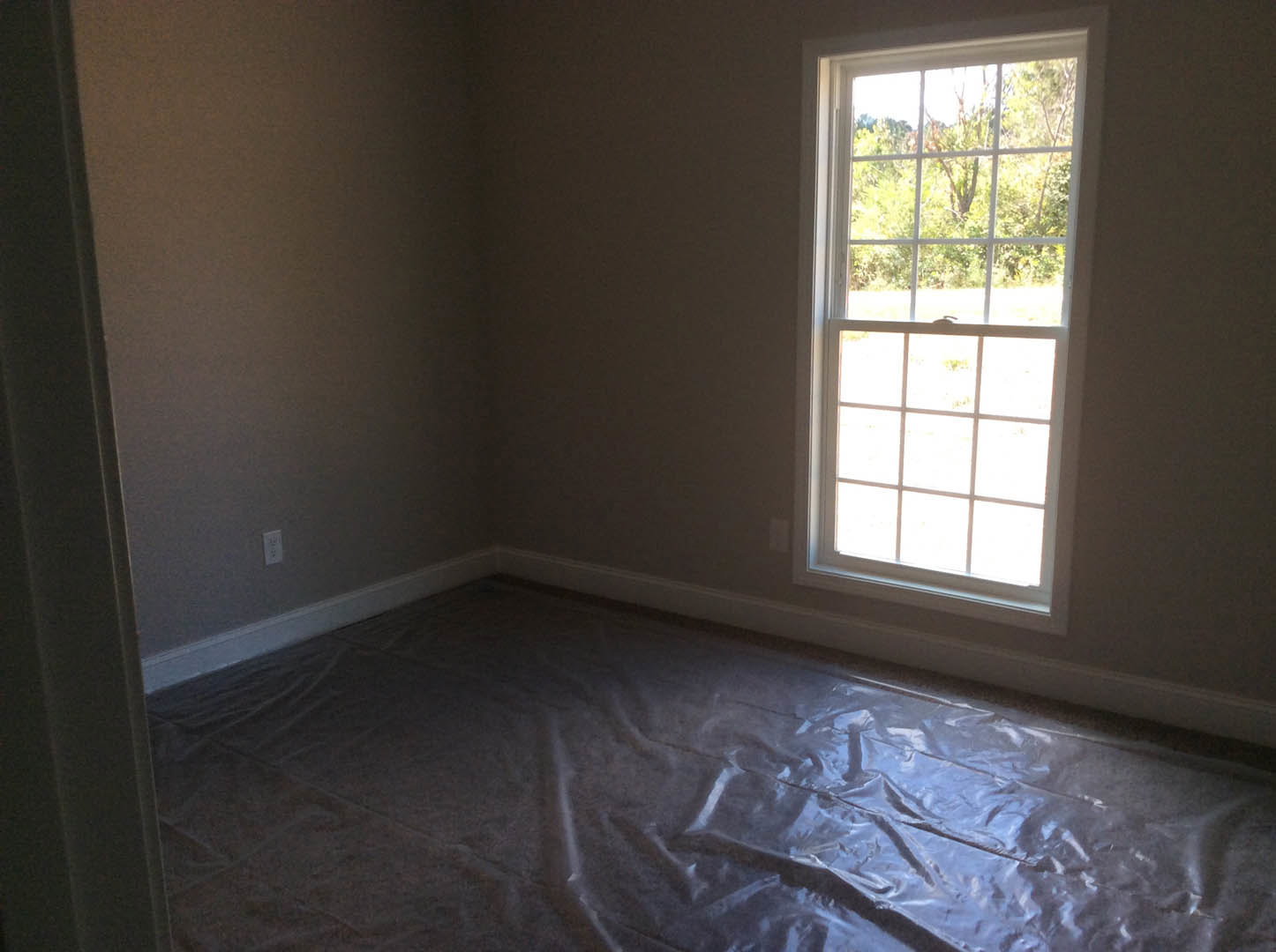 Unfinished room with multi-pane window, clear plastic sheeting covering the floor, white plaster walls, and view of leafy trees outside