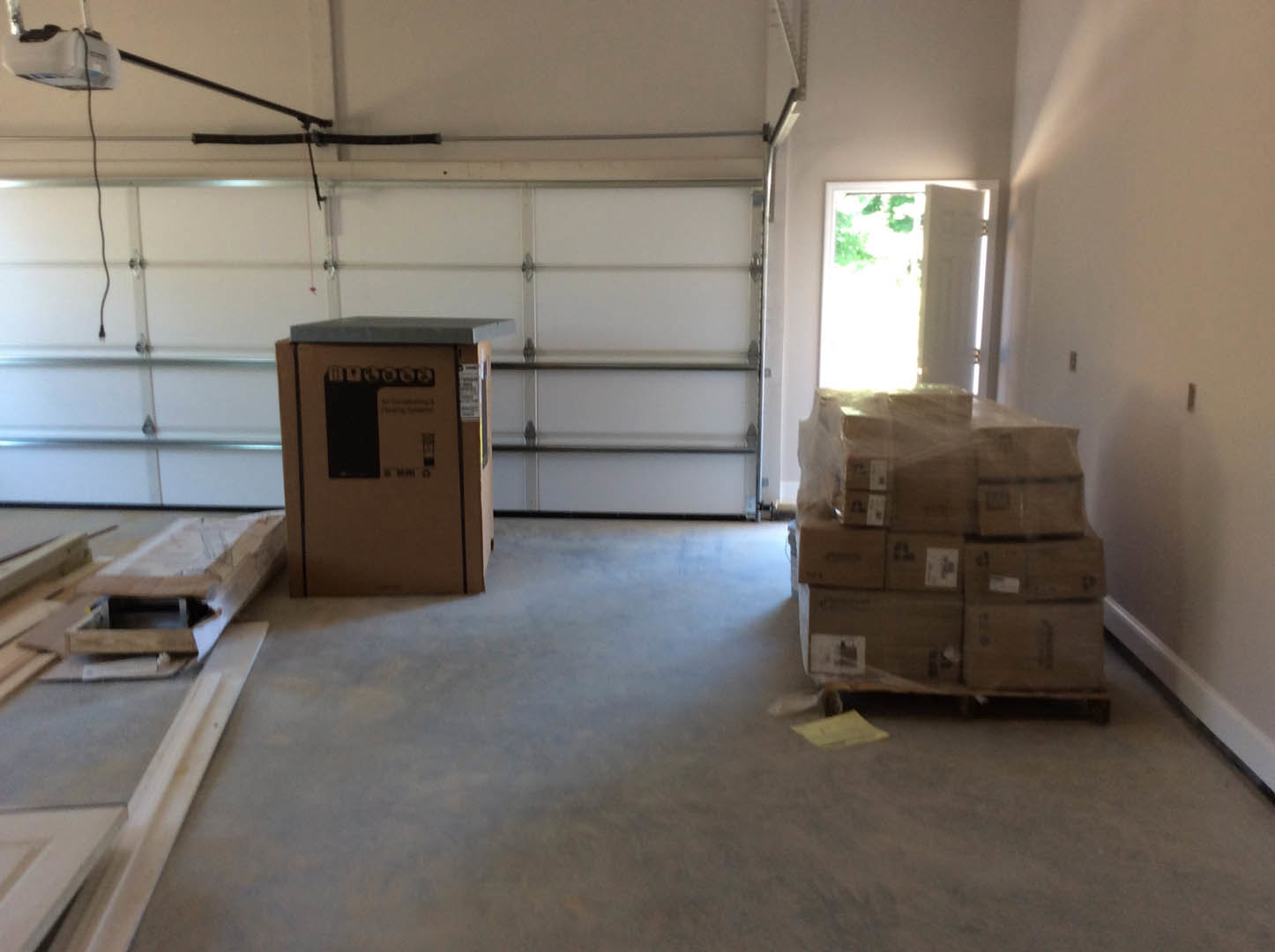 Unfinished room with stacked cardboard boxes and wooden pallets on light wood flooring, white door letting in natural light, plain white walls and ceiling