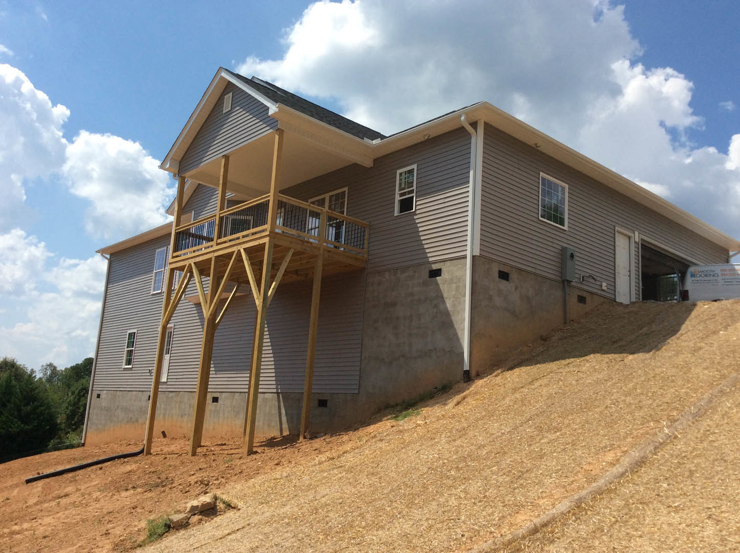 Modern cottage-style home with multiple square windows, balcony, and sloped roof, situated on a grassy hill overlooking dense trees under a cloudy sky