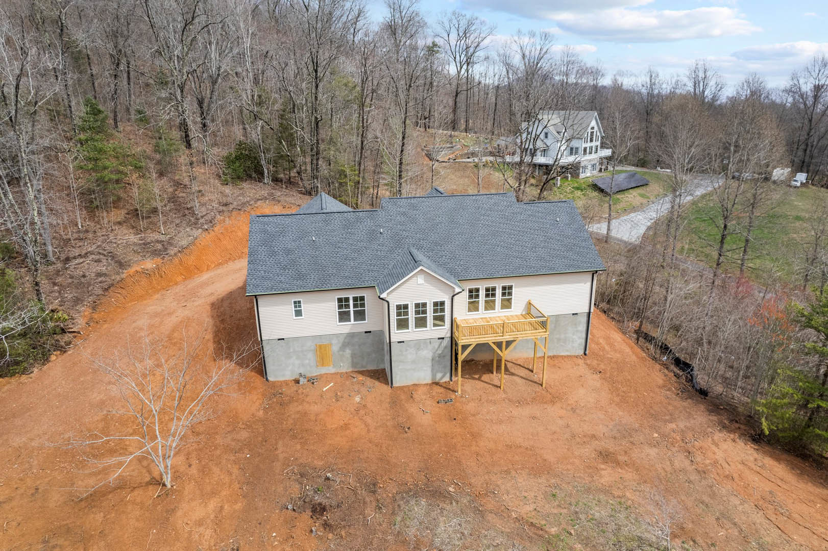 Wooden deck attached to a cottage-style house, dirt road leading up to the entrance, trees and blue sky with scattered clouds in the background, grey tarp spread on grassy area.